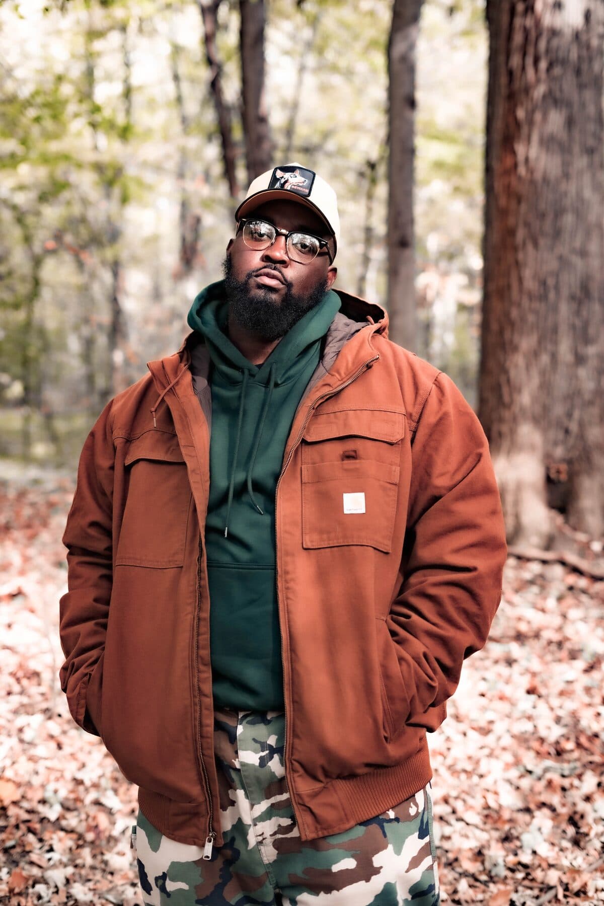 Nappi Devi stands in an autumn woodland wearing a rust jacket, forest-green hoodie, camouflage trousers, glasses and a cap.