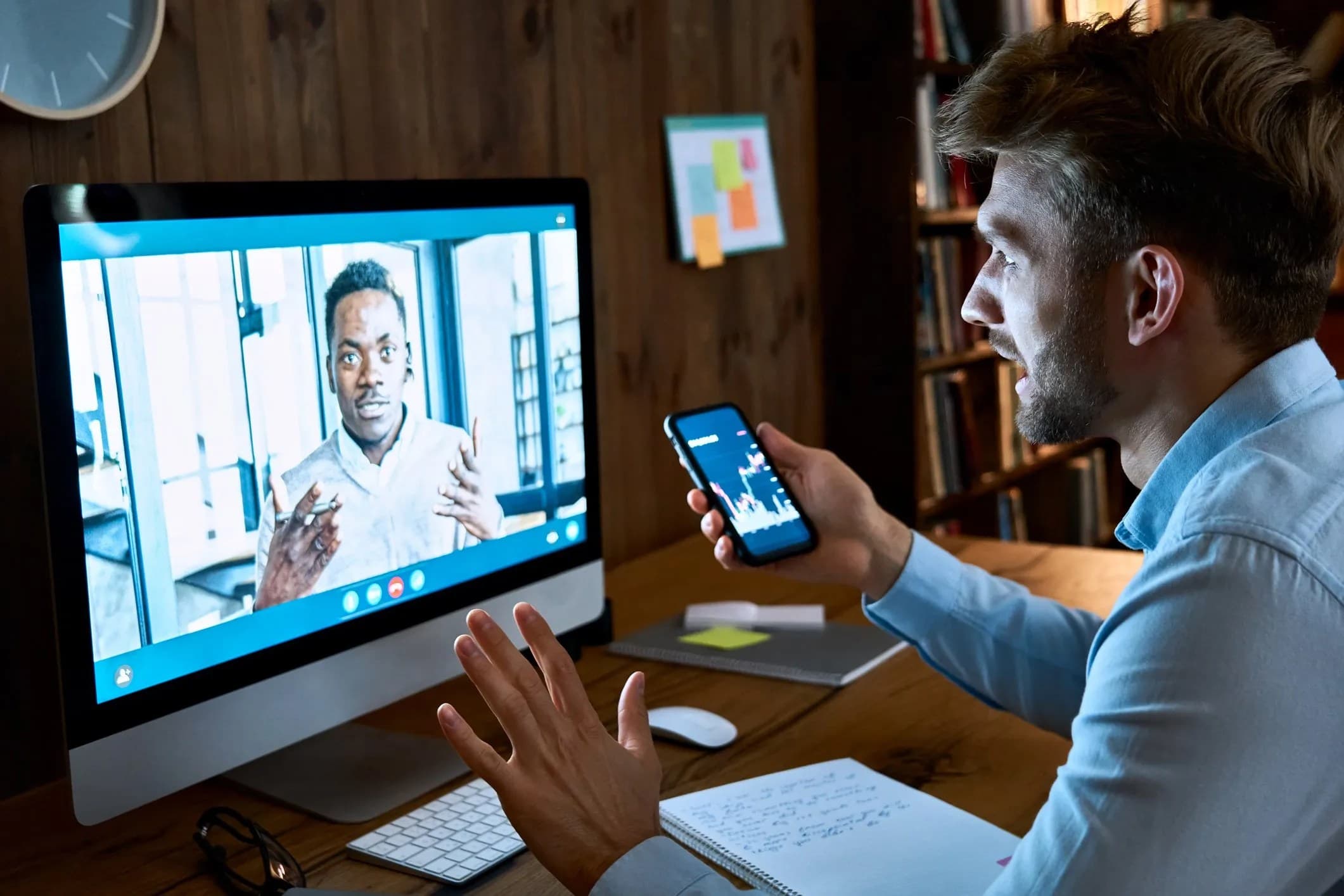 A white man sitting on a work table teleconferencing with a Black man on a Mac while holding a phone with the a stocks app open