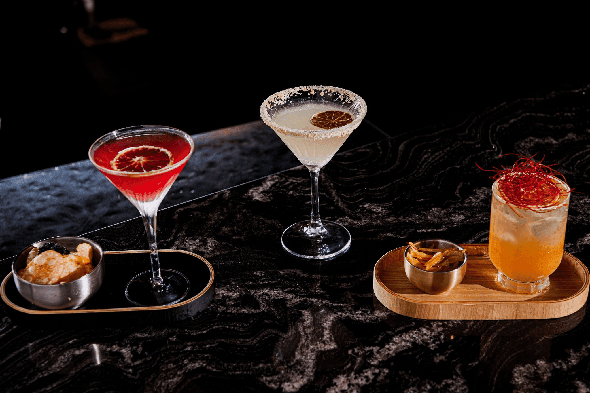 Three colorful seasonal cocktails arranged on a dark bar at Winsome Prime